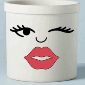 May include: A white ceramic planter with a whimsical face design. The design features a winking eye with long lashes, a curved nose, and red lips. The planter has a cylindrical shape with a slightly flared rim.