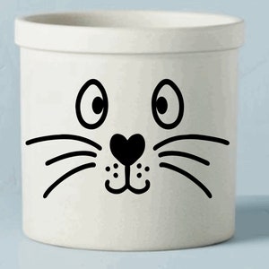 May include: A white ceramic container with a whimsical cat face design. The cat face features large, round eyes, a heart-shaped nose, and whiskers. The container is cylindrical with a slightly flared rim, suitable for kitchen or home decor.