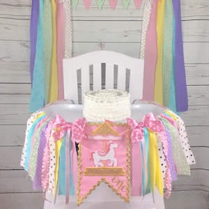 May include: A pink and gold high chair banner with a carousel horse design and the word "one" embroidered on it. The banner is decorated with pastel colored ribbons and bows. A white cake sits on the high chair.