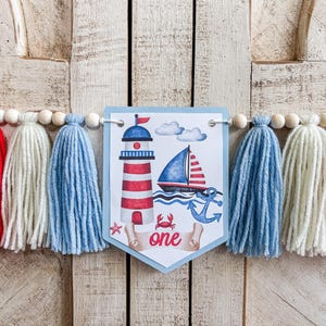 May include: A nautical-themed banner with blue, red, and white tassels. The banner features a central pennant with a lighthouse, sailboat, and anchor illustration. The word "one" is displayed on the pennant.