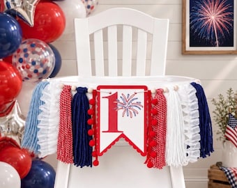 May include: A high chair banner featuring a white pennant with a red number one and a firework graphic. The banner is decorated with red, white, and blue yarn tassels, ruffled fabric strips, and red pom-poms strung on a cord with wooden beads.