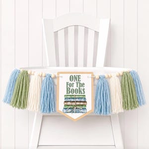 May include: A banner with blue, green, and cream tassels, wooden beads, and a sign that reads "ONE For The Books." The sign features a watercolor illustration of stacked books tied with twine. The banner is displayed on a white high chair.