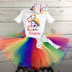 May include: Rainbow tutu with a white shirt featuring a colorful number one with a rainbow and sun graphic. The shirt is personalized with the name "Emeree".