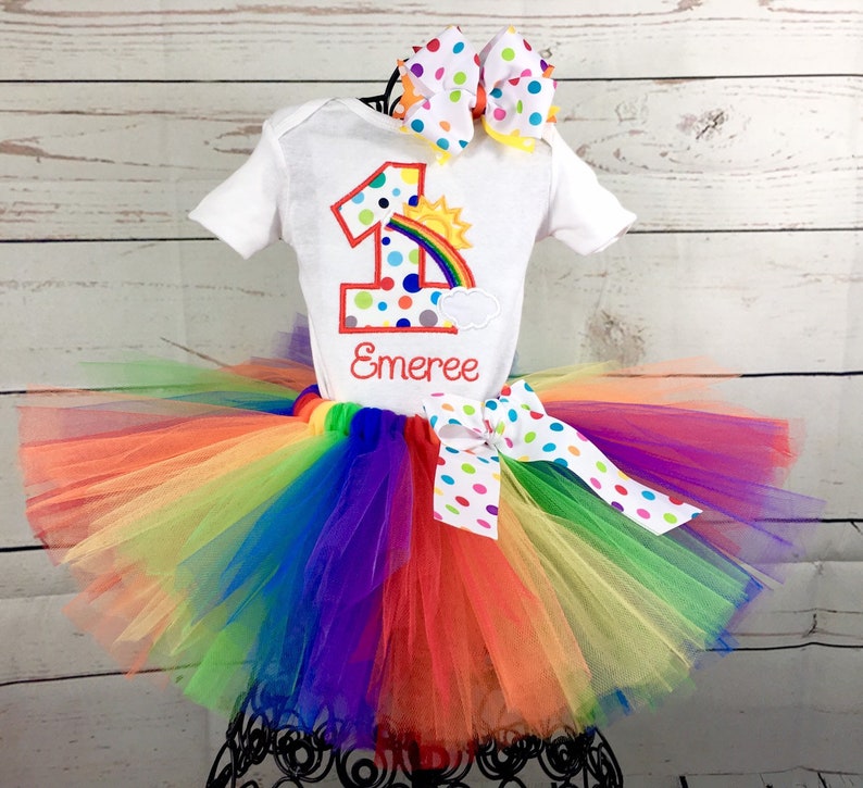 May include: Rainbow tutu with a white shirt featuring a colorful number one with a rainbow and sun graphic. The shirt is personalized with the name "Emeree".