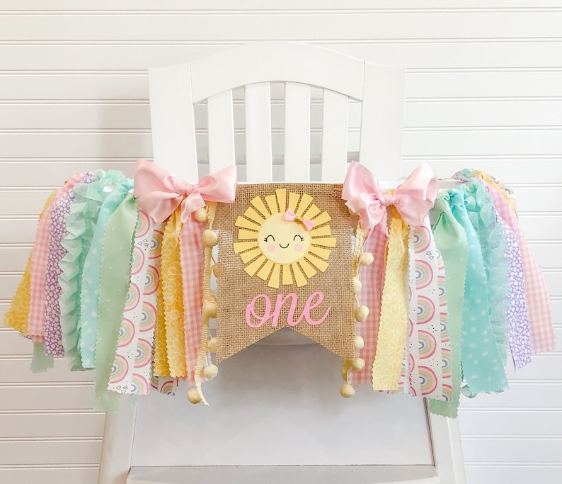 Puede incluir: Una pancarta de arpillera con un sol sonriente y la palabra "uno" en letras rosas. La pancarta est&aacute; decorada con lazos rosas y serpentinas de tela de colores arco&iacute;ris.