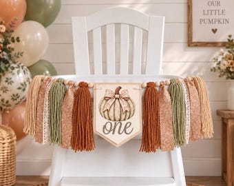 Op de afbeelding: Een kinderstoelbanner met een centraal wimpel voorzien van een pompoenillustratie en het woord one in cursief schrift. De banner is versierd met afwisselende strengen van beige, roestoranje en saliegroene garenkwastjes en stroken stof.