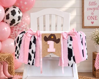May include: A high chair banner featuring a burlap pennant with a brown cowboy hat and the number one in light pink. The banner is decorated with strips of fabric in black and white cow print, light pink gingham, and solid light pink ribbons.