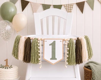 May include: A high chair banner featuring a central white pennant with the number 1 in sage green, flanked by yarn tassels in shades of olive, dark green, and cream, accented with wooden beads and small white pom-poms on a string.