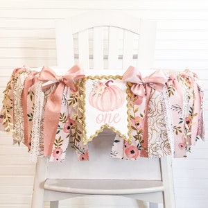 May include: A pink and gold high chair banner with a watercolor pumpkin and the word "one" in gold glitter. The banner is decorated with pink and gold ribbons, lace, and floral fabric.