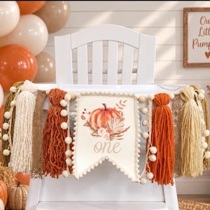 May include: A high chair banner featuring a central cream-colored pennant with a pumpkin illustration and the word one in script. The banner is decorated with orange, cream, and brown yarn tassels, lace trim, and a string of small white pom-poms.