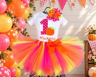 May include: A white short-sleeved bodysuit featuring a pink polka-dot number one, an orange pumpkin graphic, and the name Mackenna in cursive script, paired with a multi-layered neon pink, orange, and yellow tulle tutu skirt with a bow.