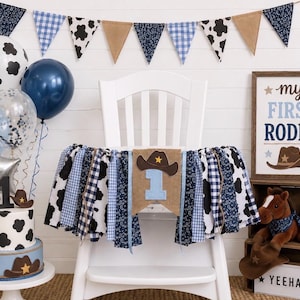 May include: A high chair banner with blue, white, and black fabric strips, featuring a burlap center panel with a brown cowboy hat and the number one. Includes balloons, a cake with a number one topper, and a sign reading My First Rodeo.