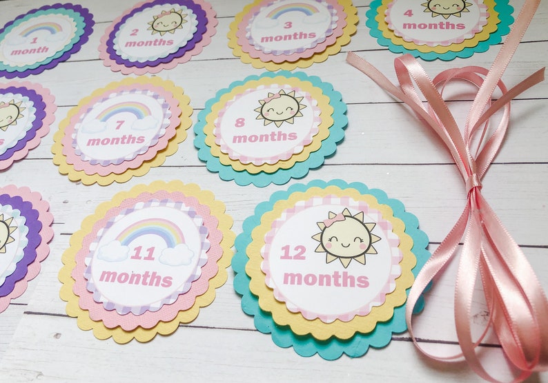 Puede incluir: Doce c&iacute;rculos de papel coloridos y festoneados con un dise&ntilde;o de arco&iacute;ris y sol. Cada c&iacute;rculo tiene un n&uacute;mero del 1 al 12 y la palabra "meses" impresa en &eacute;l. Los c&iacute;rculos est&aacute;n dispuestos en forma de semic&iacute;rculo sobre una superficie de madera. Una cinta rosa est&aacute; en el fondo.