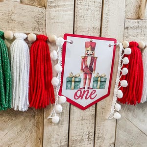 May include: A festive banner with red, green, and white yarn tassels and wooden beads. A central pennant features a Nutcracker illustration and the word "one". White pom-pom trim adds a decorative touch. Perfect for a birthday.