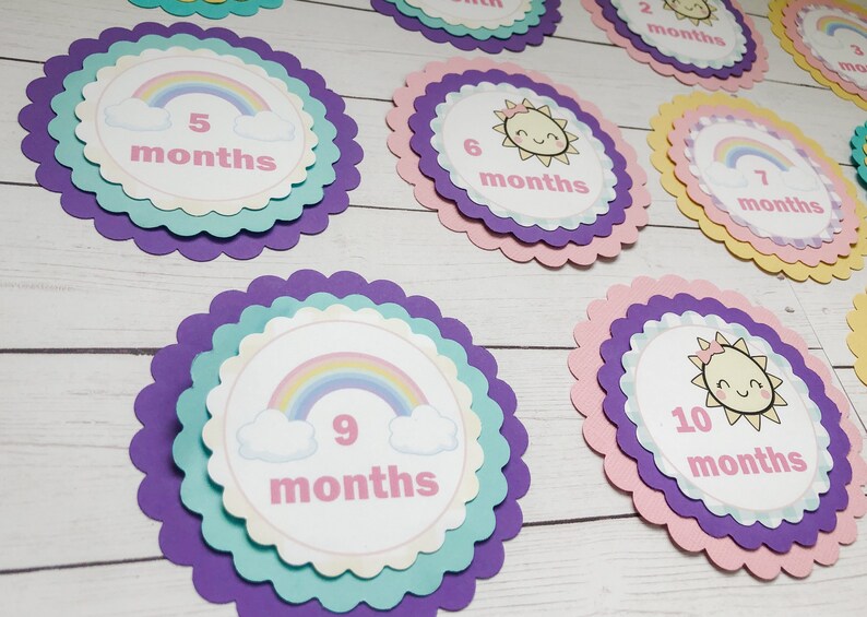Puede incluir: Un conjunto de diez tarjetas de hitos para beb&eacute;s coloridas con dise&ntilde;os de arco&iacute;ris y sol. Cada tarjeta presenta un n&uacute;mero del 1 al 10 y la palabra "meses".