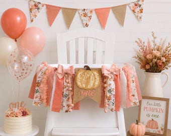 May include: A high chair banner featuring a burlap pennant with a gold sequin pumpkin and the word one in script, surrounded by fabric strips in peach, white, and floral patterns with bows, displayed on a white high chair with balloons.