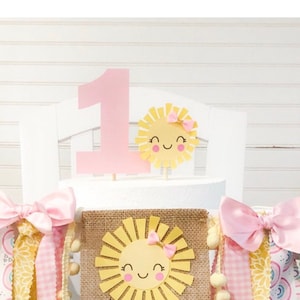 Puede incluir: Un topper de pastel de color rosa con el n&uacute;mero uno, con un sol amarillo con un lazo rosa y una cara sonriente. Un banner de arpillera con un sol amarillo con un lazo rosa y una cara sonriente. El banner tiene cintas rosas y amarillas.