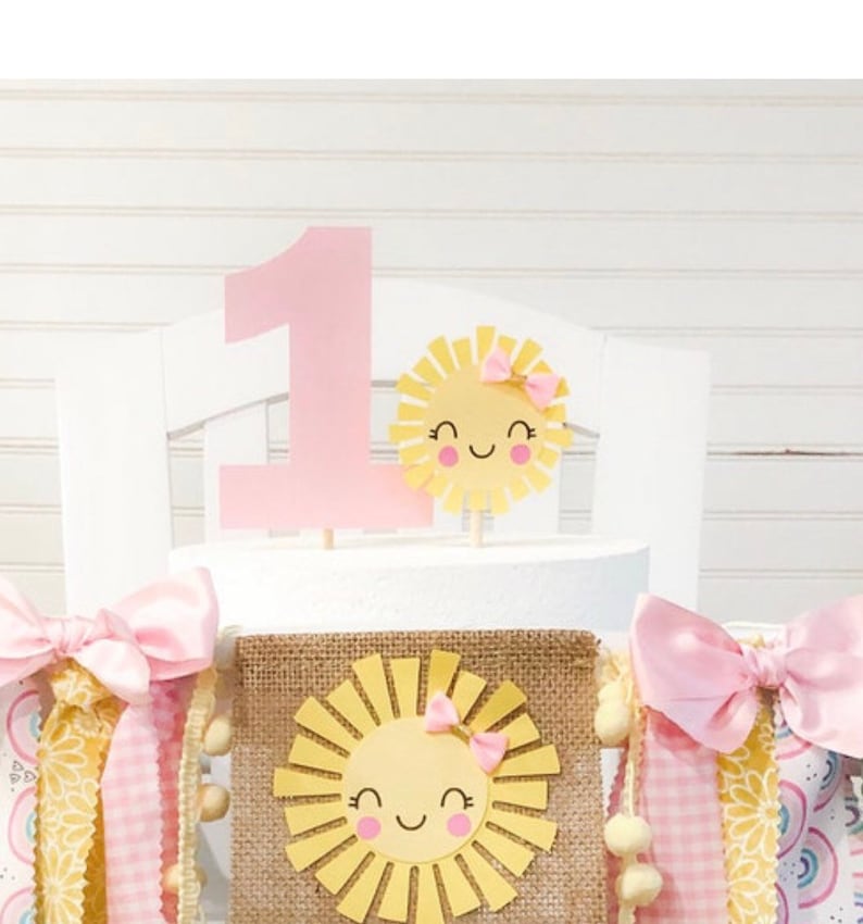 Puede incluir: Un topper de pastel de color rosa con el n&uacute;mero uno, con un sol amarillo con un lazo rosa y una cara sonriente. Un banner de arpillera con un sol amarillo con un lazo rosa y una cara sonriente. El banner tiene cintas rosas y amarillas.
