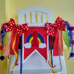 May include: A red and white high chair banner with a rainbow and the number one on it. The banner has white polka dots and white pom poms on the edges. There are red and white polka dot bows on each end of the banner. There are colorful ribbons hanging from the banner.