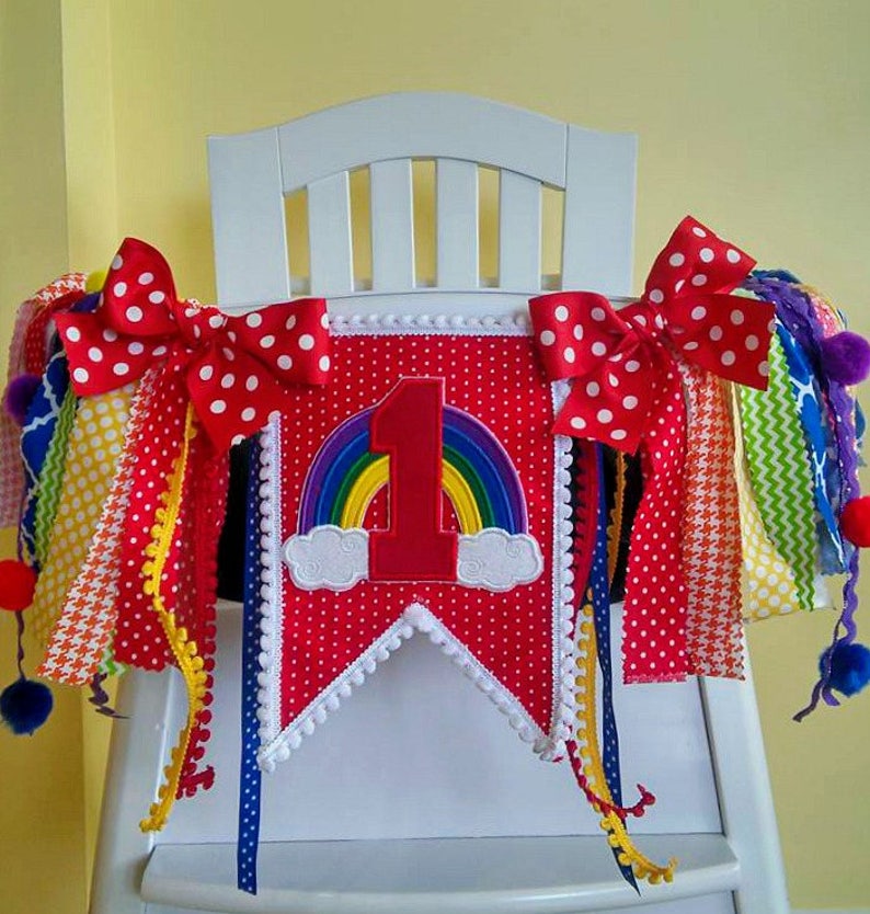 May include: A red and white high chair banner with a rainbow and the number one on it. The banner has white polka dots and white pom poms on the edges. There are red and white polka dot bows on each end of the banner. There are colorful ribbons hanging from the banner.