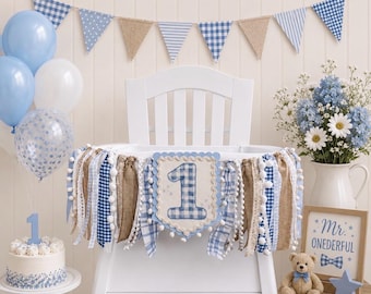 May include: A high chair banner featuring a central square with the number 1 in blue gingham, surrounded by hanging fabric strips in blue, white, and tan. The banner includes lace, pom-pom trim, and burlap accents, creating a decorative display for a first birthday celebration.