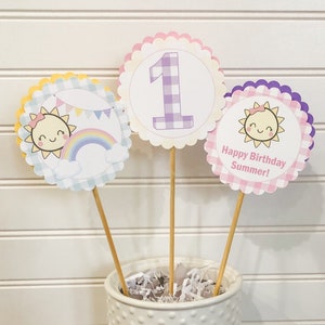 Puede incluir: Tres toppers de cupcake de papel de color pastel con un patr&oacute;n de cuadros. Los toppers presentan un sol sonriente con un arco&iacute;ris, el n&uacute;mero uno y el texto "&iexcl;Feliz cumplea&ntilde;os de verano!"