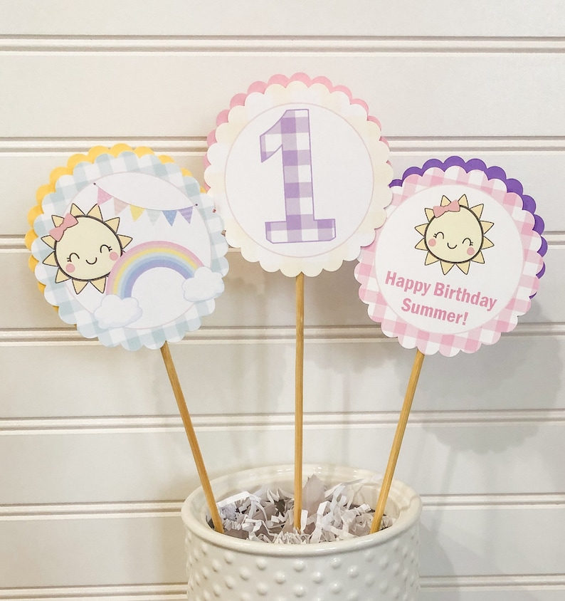 Puede incluir: Tres toppers de cupcake de papel de color pastel con un patr&oacute;n de cuadros. Los toppers presentan un sol sonriente con un arco&iacute;ris, el n&uacute;mero uno y el texto "&iexcl;Feliz cumplea&ntilde;os de verano!"