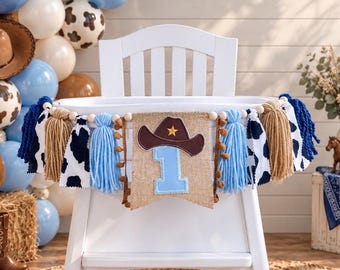 May include: A high chair banner featuring a central burlap pennant with a light blue number one and a brown cowboy hat applique. The banner is decorated with light blue and tan yarn tassels, white and black cow print fabric, and wooden beads.
