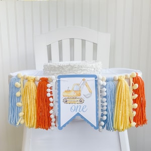 May include: A blue and yellow high chair banner with a yellow excavator and the word "one" on it. The banner is decorated with blue, yellow, and orange tassels and white pom-poms.