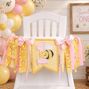 May include: A yellow and white polka dot fabric banner featuring a large pink number one and a smiling bee appliqu&eacute;. The banner is decorated with pink satin bows and hanging fabric strips in yellow and pink, accented with small pom-poms.