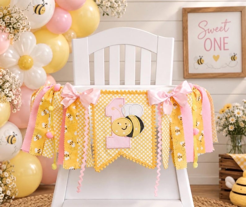 May include: A yellow and white polka dot fabric banner featuring a large pink number one and a smiling bee appliqu&eacute;. The banner is decorated with pink satin bows and hanging fabric strips in yellow and pink, accented with small pom-poms.
