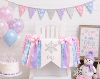 May include: A high chair banner featuring a white fabric pennant with a silver glitter snowflake and the word one, decorated with pink, blue, and purple ribbons and bows. The banner is displayed on a white chair with balloons and a sign.