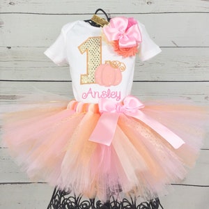 May include: A pink and orange tulle tutu with a pink satin bow. A white onesie with a gold glitter number one and a pumpkin design. The onesie is personalized with the name "Ansley".