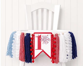 May include: A festive high chair banner with red, white, and blue tassels and pom-poms. The banner features a white pennant with a red number 1 and a firework design, perfect for a first birthday celebration.