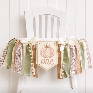 May include: A high chair banner featuring a central pennant with a pumpkin illustration and the word one in script. The banner is decorated with fabric strips in sage green, cream, and floral patterns, accented by two cream satin bows.