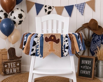May include: A white high chair decorated with a fabric banner featuring blue, brown, and cow-print strips, white pom-poms, and a central burlap pennant displaying a brown cowboy hat icon above the number 1 in a tan felt material.
