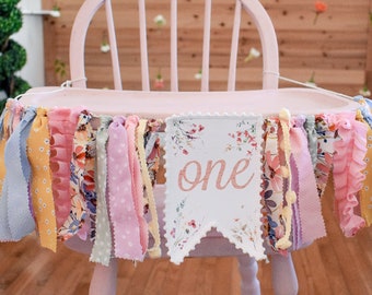 May include: A pink high chair with a white fabric banner that says "one" in gold lettering. The banner is decorated with floral designs and is surrounded by a garland of colourful fabric scraps.