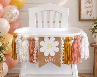May include: A high chair banner featuring a central burlap pennant with a white daisy cutout and the word one in pink script. The banner includes mustard yellow, dusty pink, and white yarn tassels, wooden beads, and daisy trim accents.