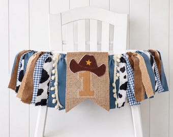 May include: A high chair banner featuring a central burlap pennant with a brown cowboy hat and the number one. The banner is decorated with strips of blue gingham, cow print, solid blue, and brown fabric, accented with white pom-pom trim.