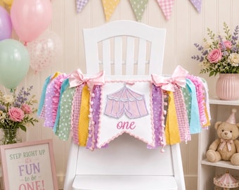 May include: A white high chair banner featuring a central white pennant with a pink circus tent graphic and the word one in pink script. The banner is decorated with multi-colored fabric strips in pastel shades of pink, yellow, green, and purple.
