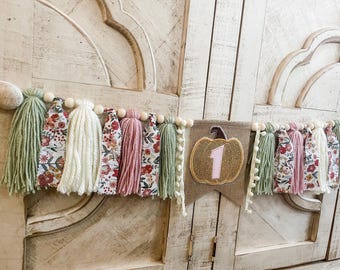 Op de afbeelding: Een banner van jute met een pompoen en het nummer één erop. De banner is versierd met groene, roze en witte kwastjes.