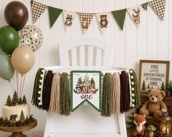 May include: A high chair banner featuring a central pennant with a bear, raccoon, and fox illustration, the word one, and thick yarn tassels in shades of green, brown, and beige. The banner is decorated with wooden beads and white pom-pom trim.