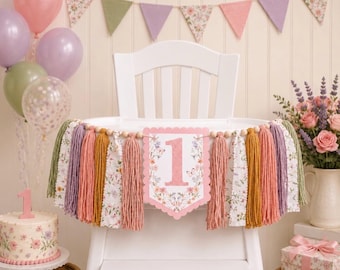 May include: A high chair banner featuring a central scalloped sign with the number 1, surrounded by floral and butterfly illustrations. The banner is decorated with alternating yarn tassels in shades of pink, mustard, and purple, and fabric strips.