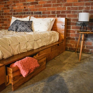 May include: A wooden bed frame with built-in drawers, a white comforter, and decorative pillows. A coral-colored blanket is visible in an open drawer. A small side table with a lamp and framed photo sits nearby, against a brick wall.