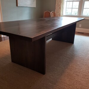 May include: A dark brown wooden dining table with a rectangular top and a unique base design. The table is made of solid wood and has a natural finish. The table is sitting on a beige rug.