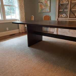 May include: A dark brown wooden dining table with a simple design and a single shelf. The table is in a room with a rug and a window.