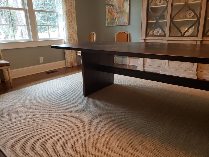 May include: A dark brown wooden dining table with a simple design and a single shelf. The table is in a room with a rug and a window.