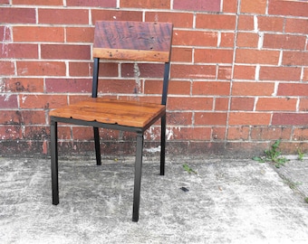 May include: A wooden chair with a black metal frame. The chair has a wide, rectangular seat and a tall, rectangular back. The chair is sitting on a concrete surface in front of a brick wall.