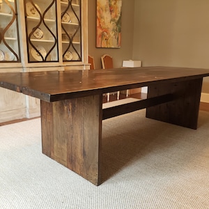 May include: A dark brown wooden dining table with a rectangular top and a simple, modern design. The table has a single shelf underneath the top, and the legs are made of thick, solid wood.