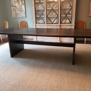 May include: A dark brown wooden dining table with a single shelf underneath. The table is in a room with a light gray wall and a rug on the floor.